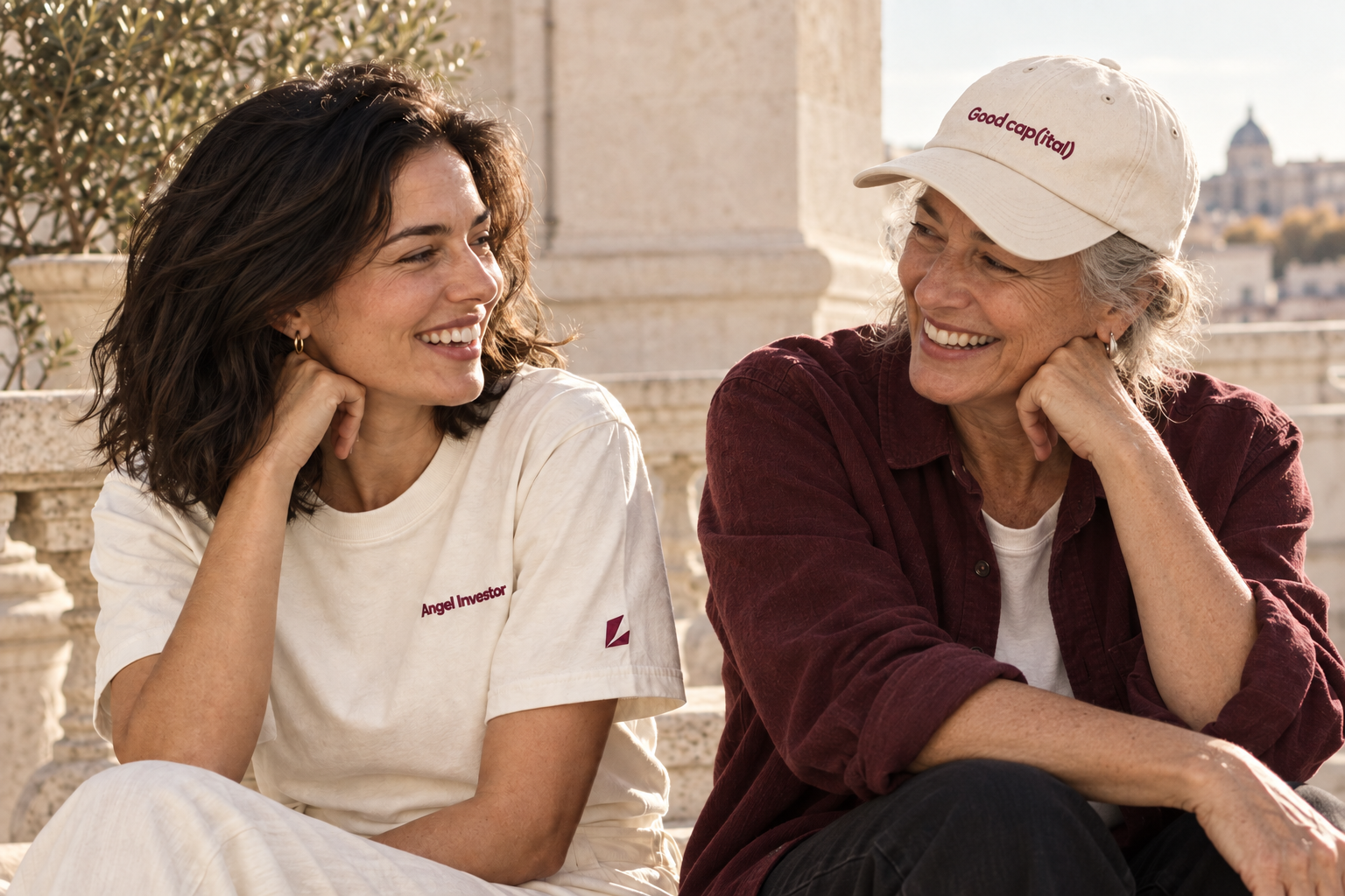 Two women on a sunlit terrace, one wearing the embroidered Angel Investor tee and the older woman wearing the embroidered Good cap(ital) cap